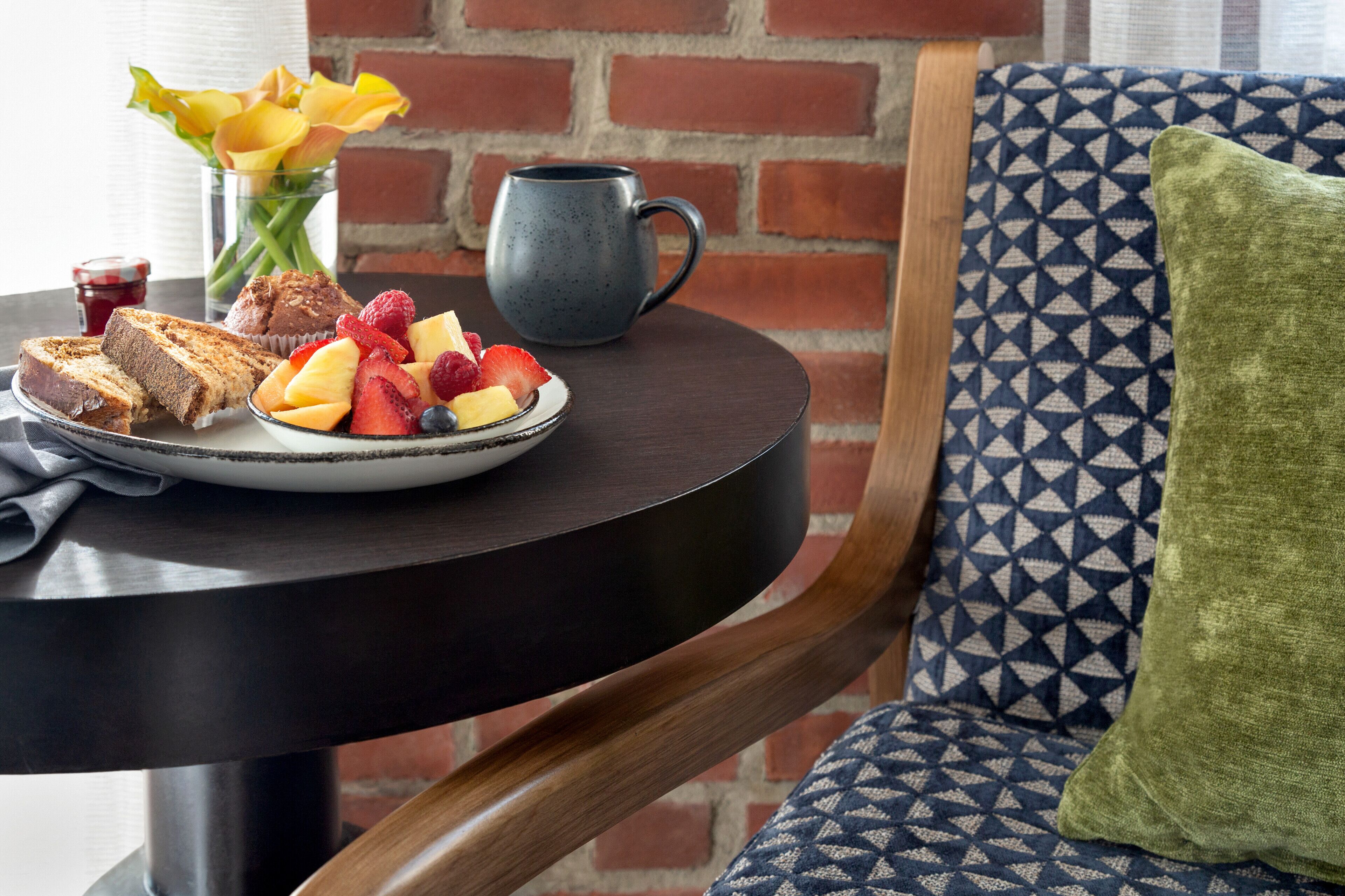 A breakfast table with toast, fruit, and a mug, set against a brick wall and patterned cushion.