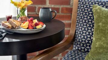 A breakfast table with toast, fruit, and a mug, set against a brick wall and patterned cushion.