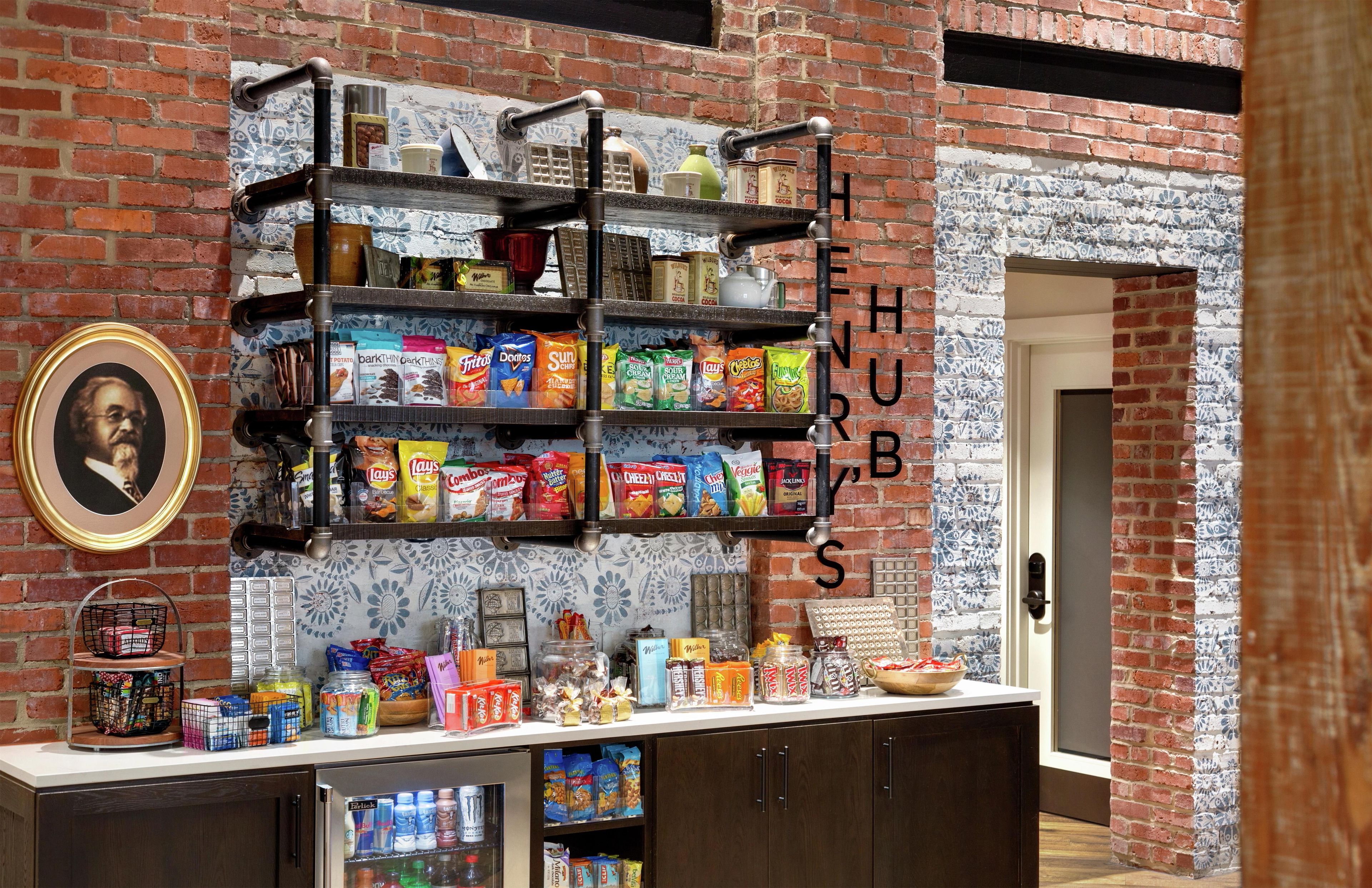 A kitchen area with a brick wall, a shelf unit with snacks, a framed picture, and a refrigerator.