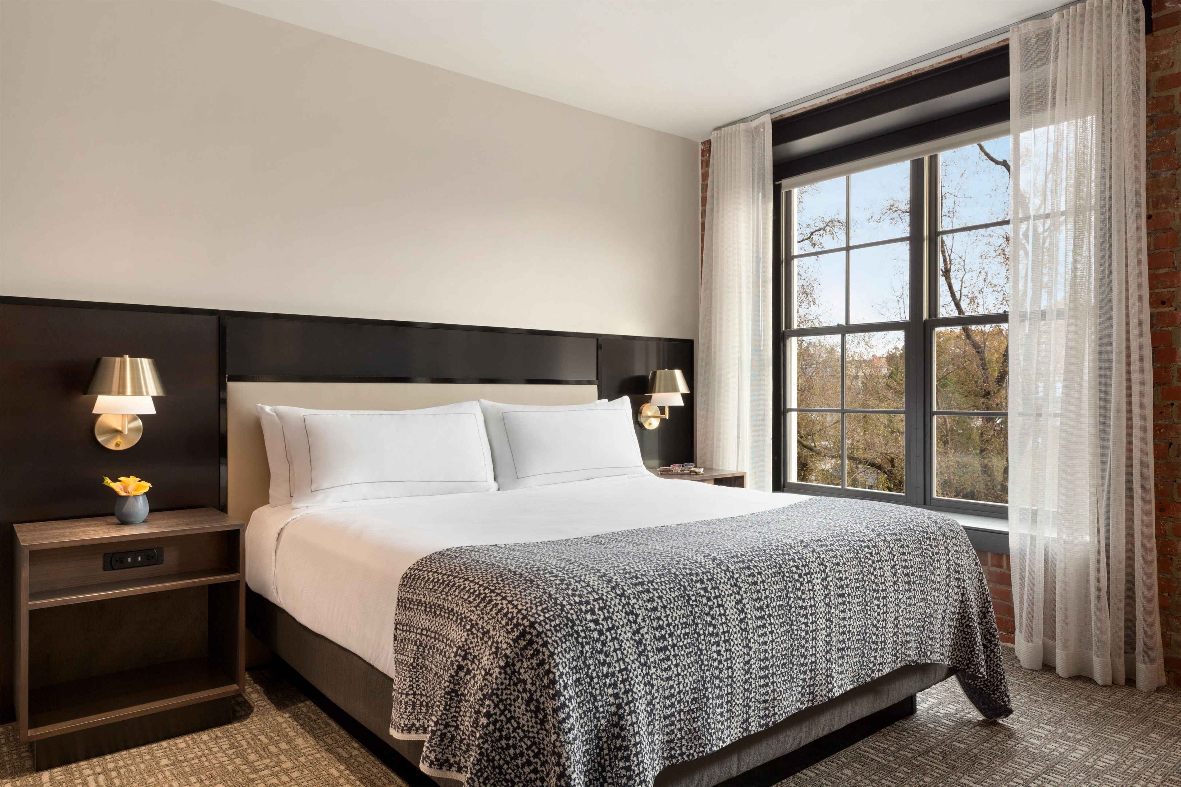 A hotel room with a large bed, two bedside tables with lamps, a view of trees through a window, and a brick wall.