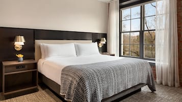 A hotel room with a large bed, two bedside tables with lamps, a view of trees through a window, and a brick wall.