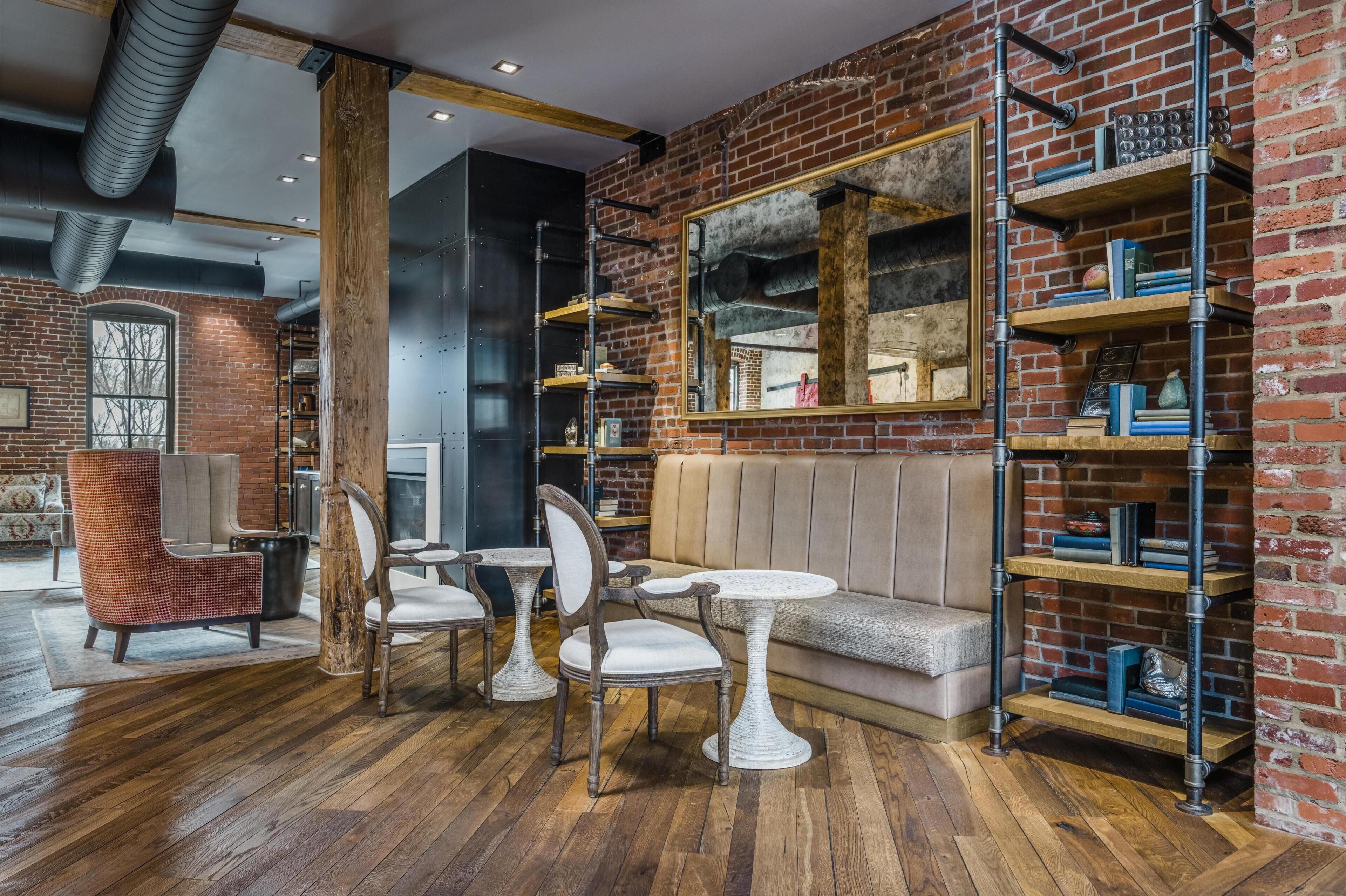 A room with a brick wall, wooden floor, a sofa, chairs, tables, and a bookshelf.
