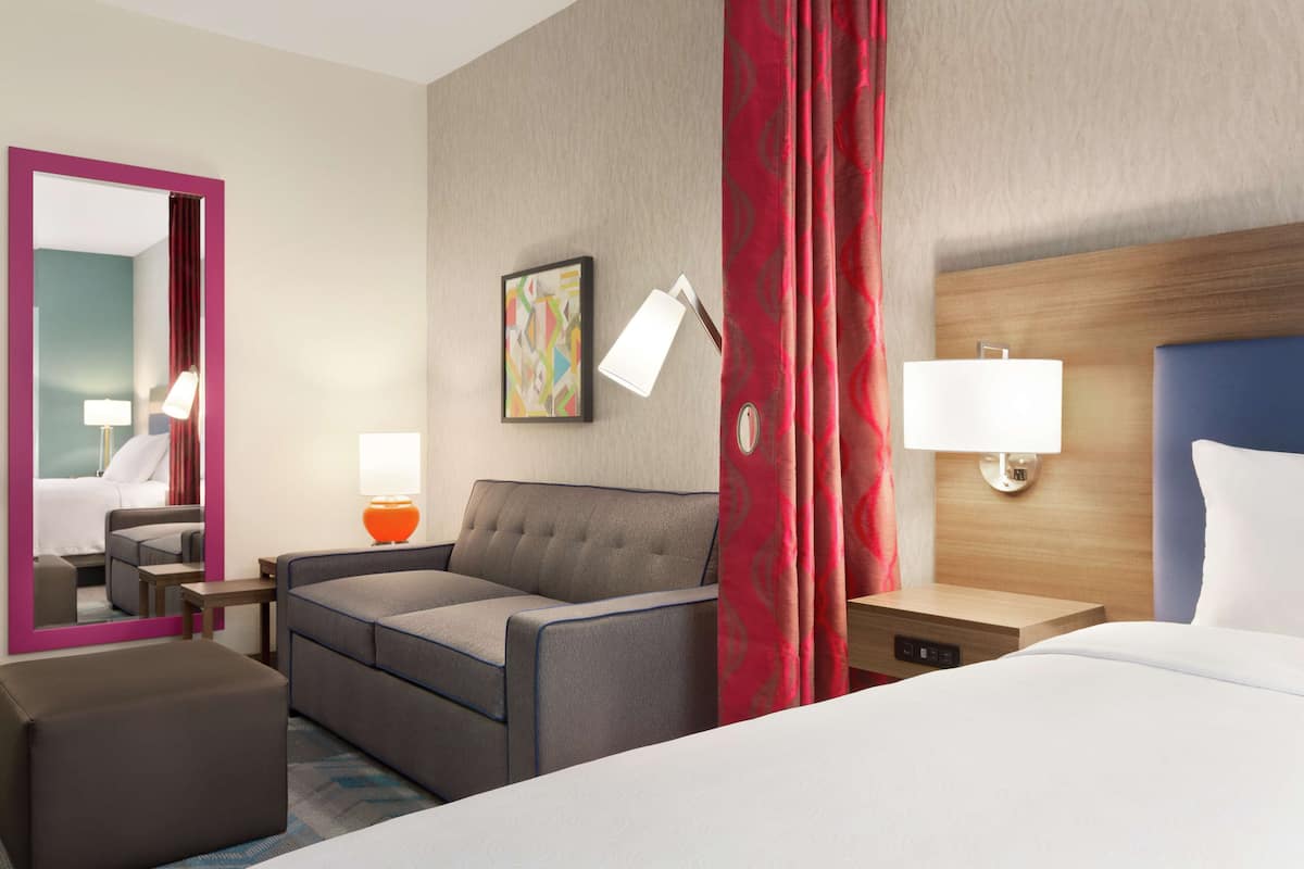 A hotel room with a bed, a sofa, a desk, a chair, a lamp, and a mirror.