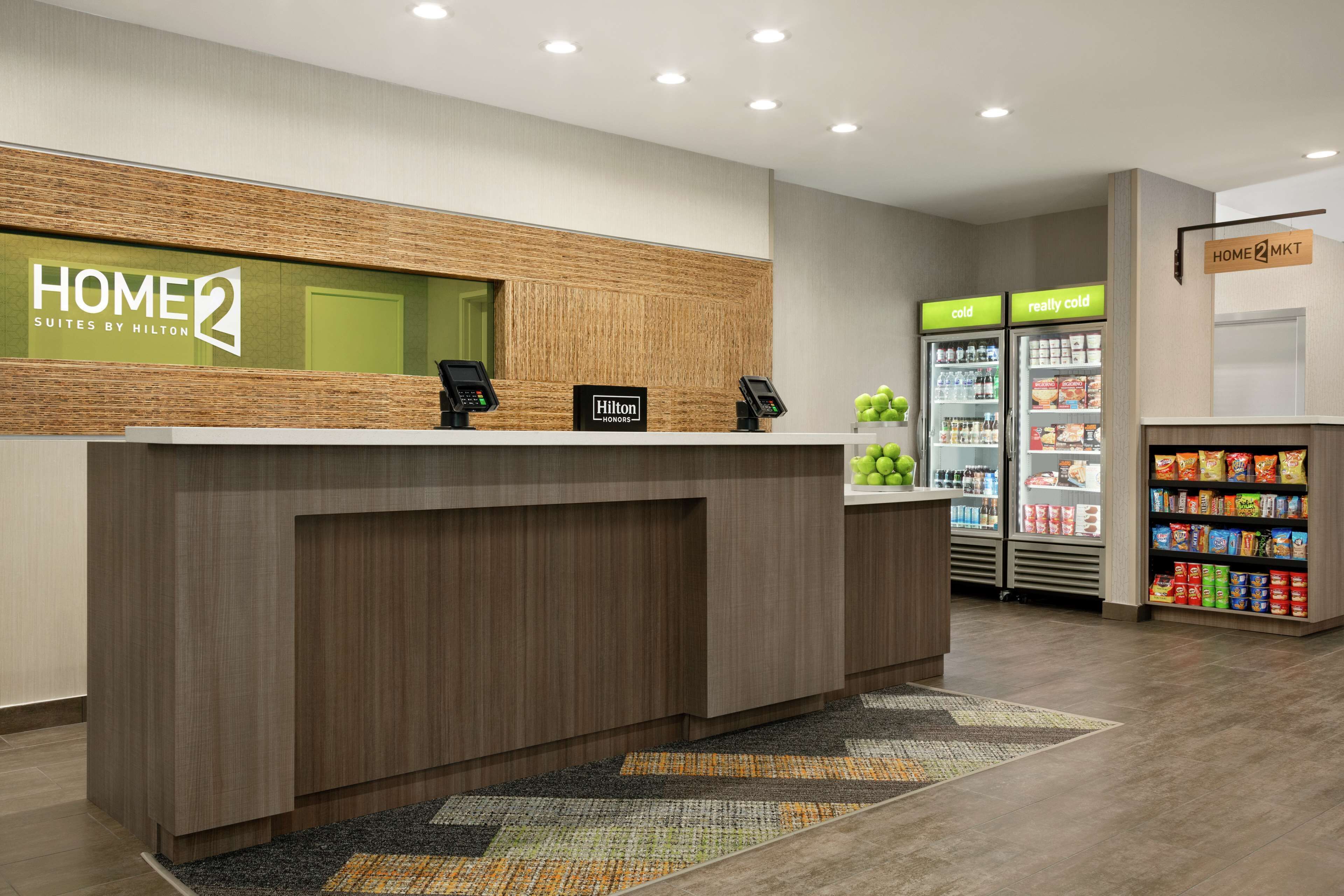 A hotel reception desk with a 'HOME 2' sign, a 'Hilton' sign, a 'cold' and 'really cold' beverage fridge, and a snack shelf.
