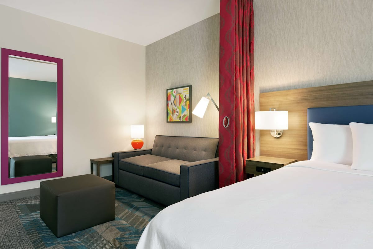 A hotel room with a bed, a sofa, a small table, a lamp, and a painting on the wall.