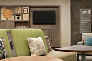 A modern living room with a sofa, a coffee table, and a flat-screen TV mounted on the wall. - Home2 Suites by Hilton Houston-Pearland (Houston)