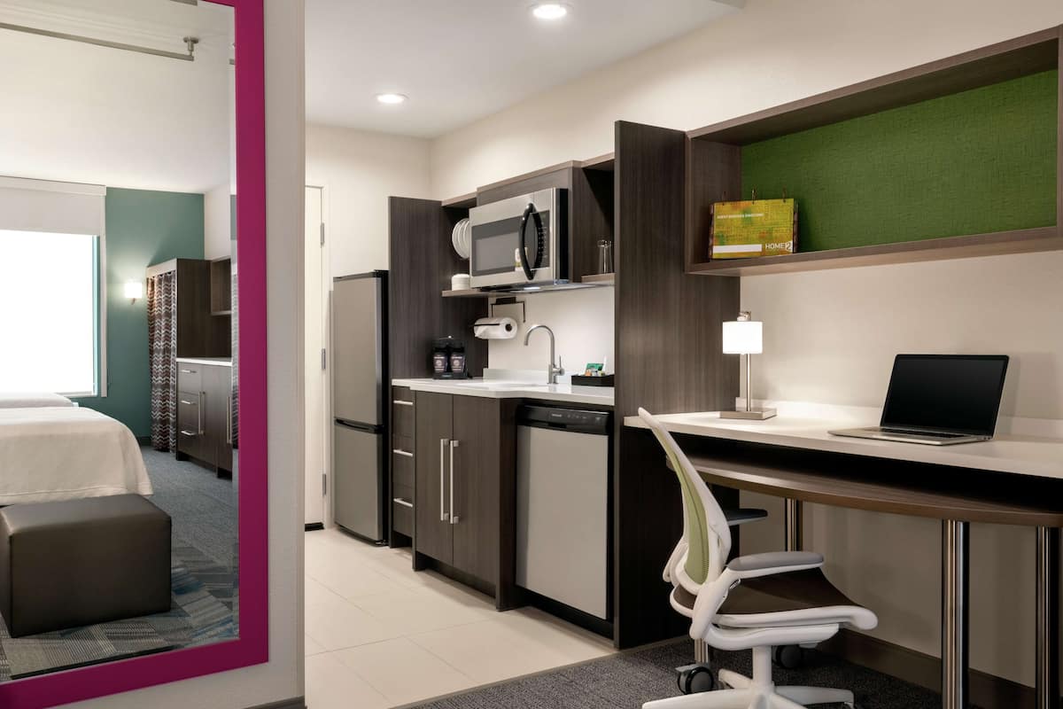 A modern hotel room with a kitchenette, a bed, a desk with a laptop, and a chair.