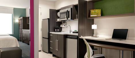 A modern hotel room with a kitchenette, a bed, a desk with a laptop, and a chair.