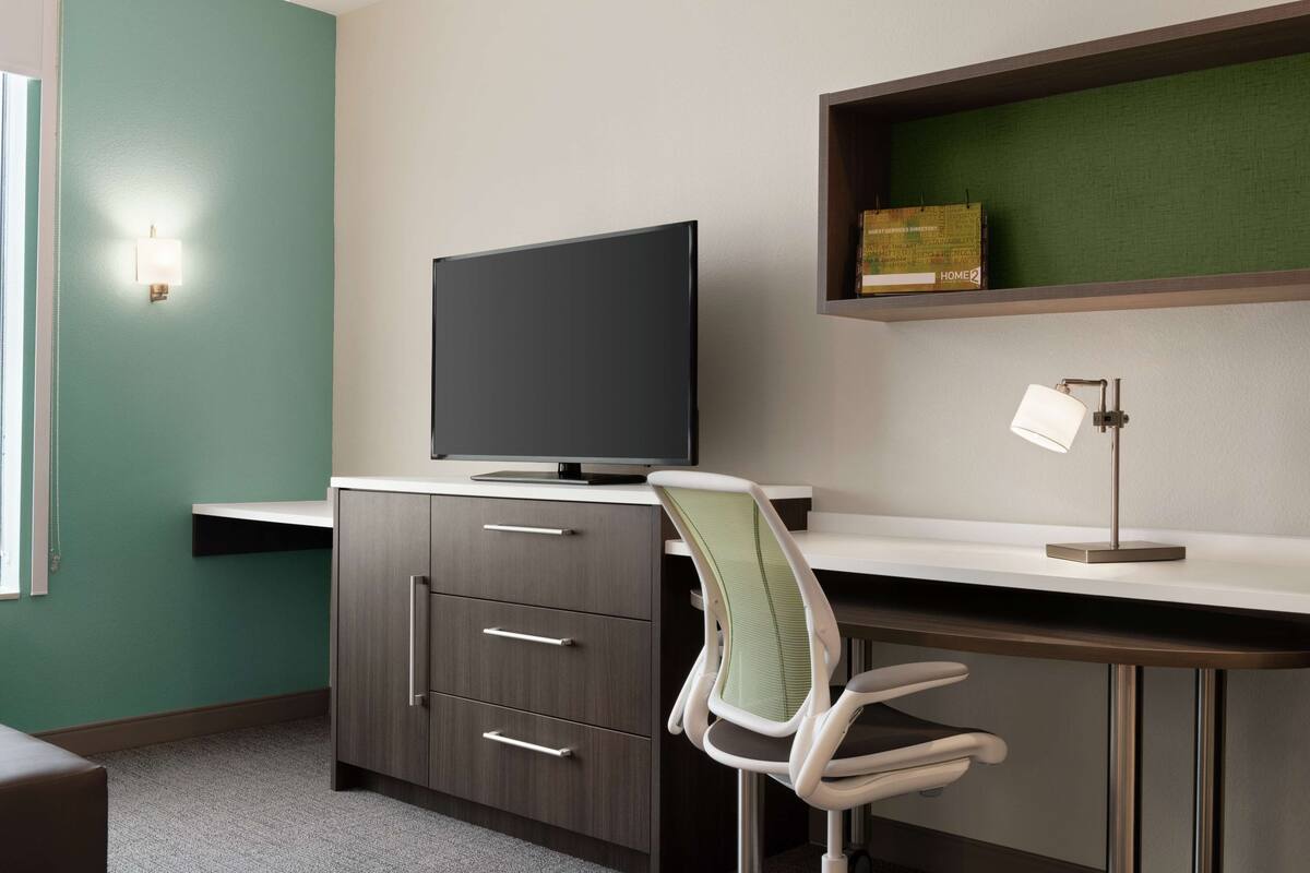 A modern office space with a flat-screen TV, a desk, a chair, a lamp, and a shelf with a box labeled "HOME2".
