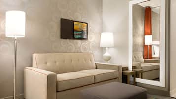 A hotel room with a sofa, a side table, a lamp, a painting, and a mirror reflecting the room.