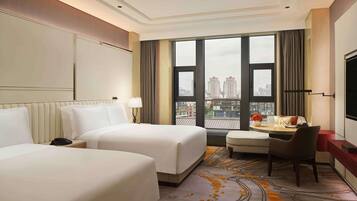 A hotel room with two beds, a desk, a chair, a TV, and a large window with a city view.