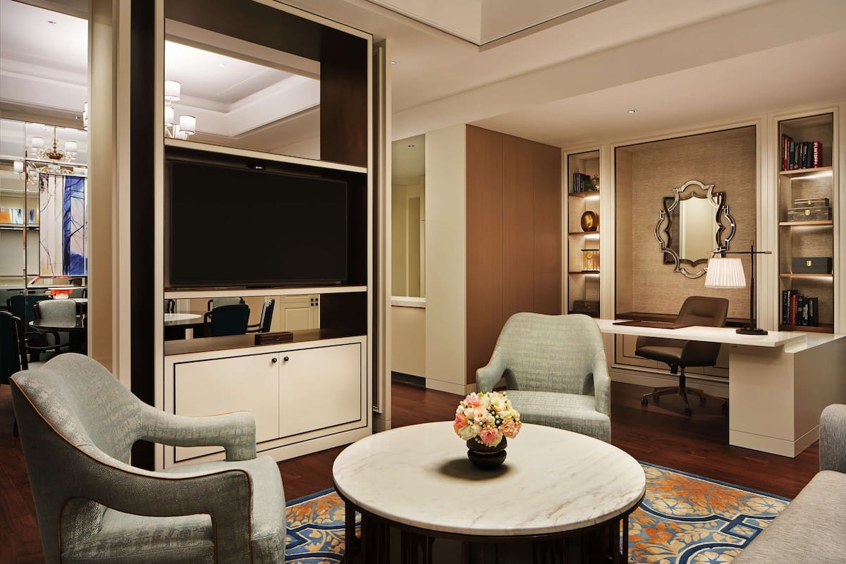 A modern hotel room with a large flat-screen TV, comfortable seating, a desk, and a bookshelf.