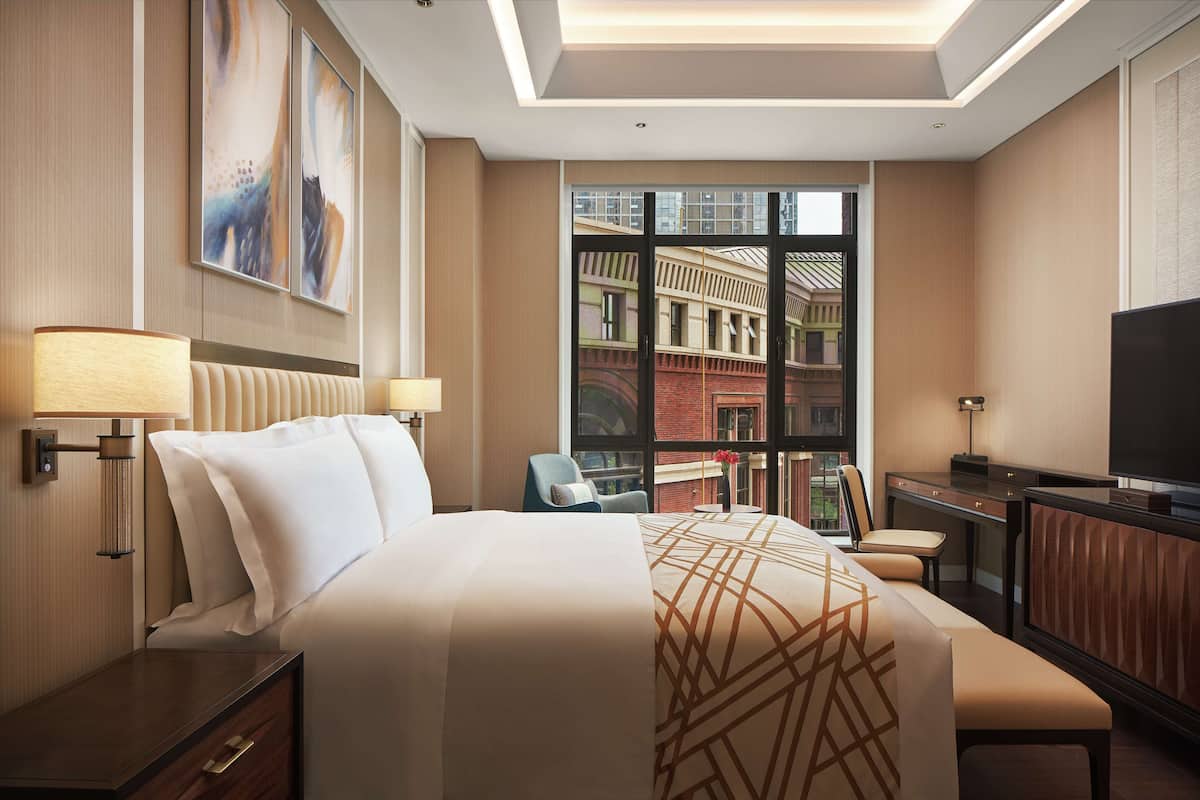 A hotel room with a large bed, a desk, a TV, and a window with a city view.