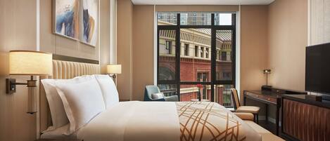 A hotel room with a large bed, a desk, a TV, and a window with a city view.