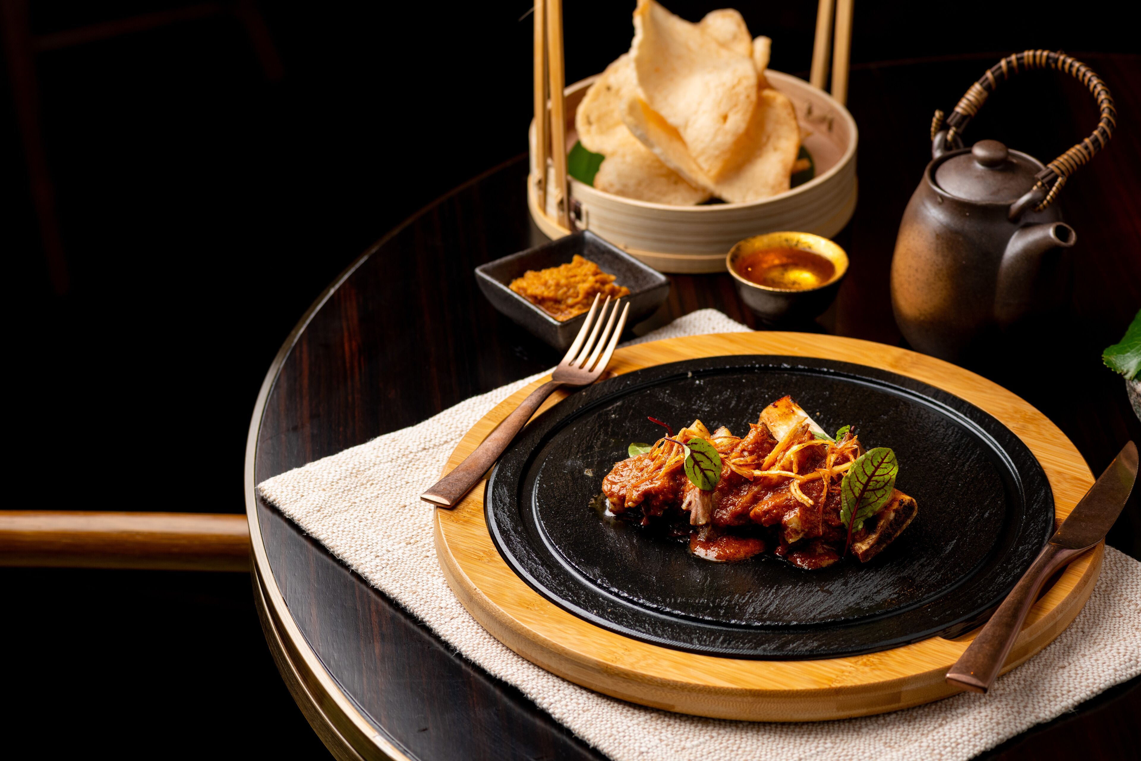 A round dining table with a black plate of food, a teapot, and a bowl of flatbread.