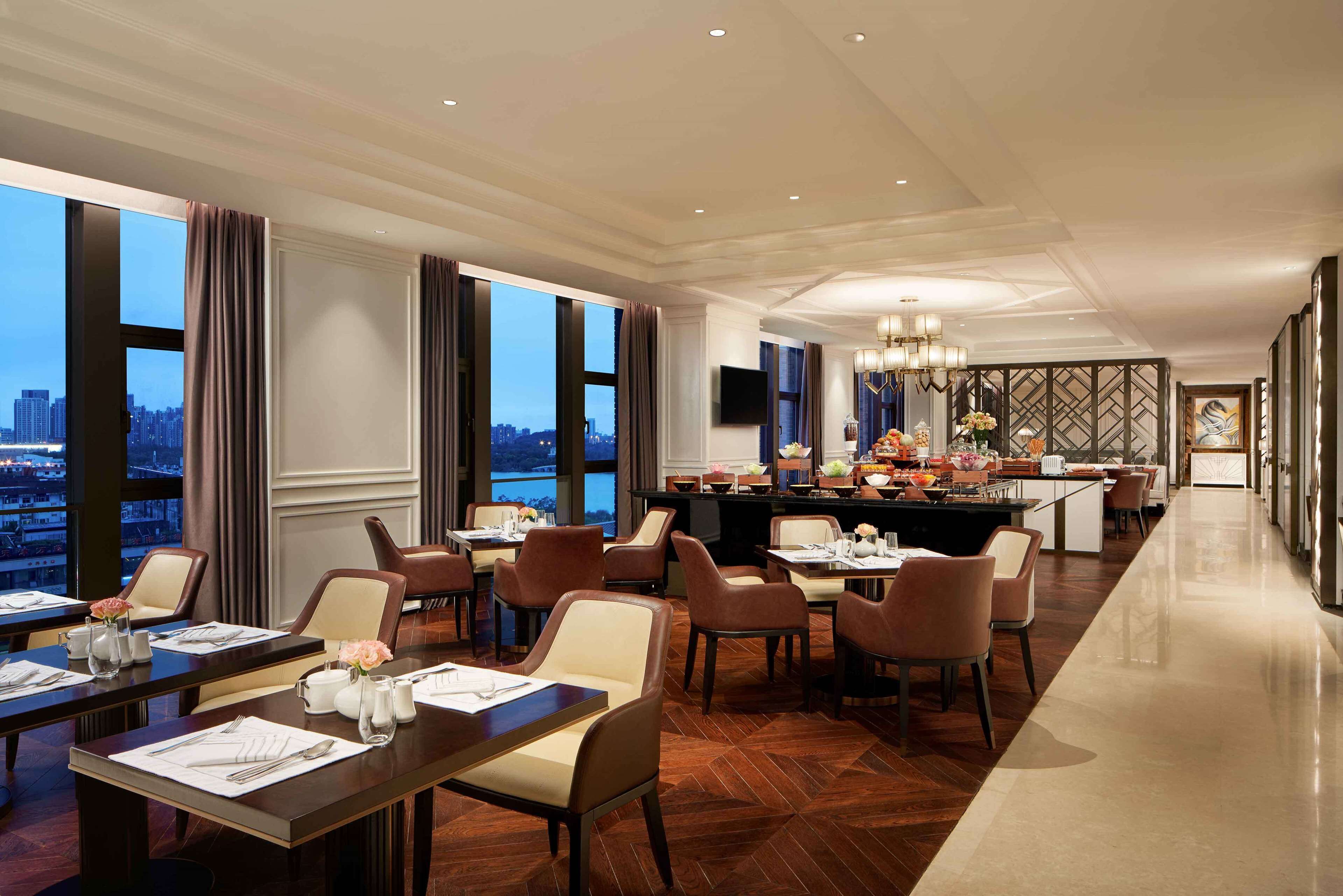 Un vaste espace de restauration dans l’hôtel, avec de nombreuses tables et chaises, un bar central et de grandes fenêtres donnant sur la ville.