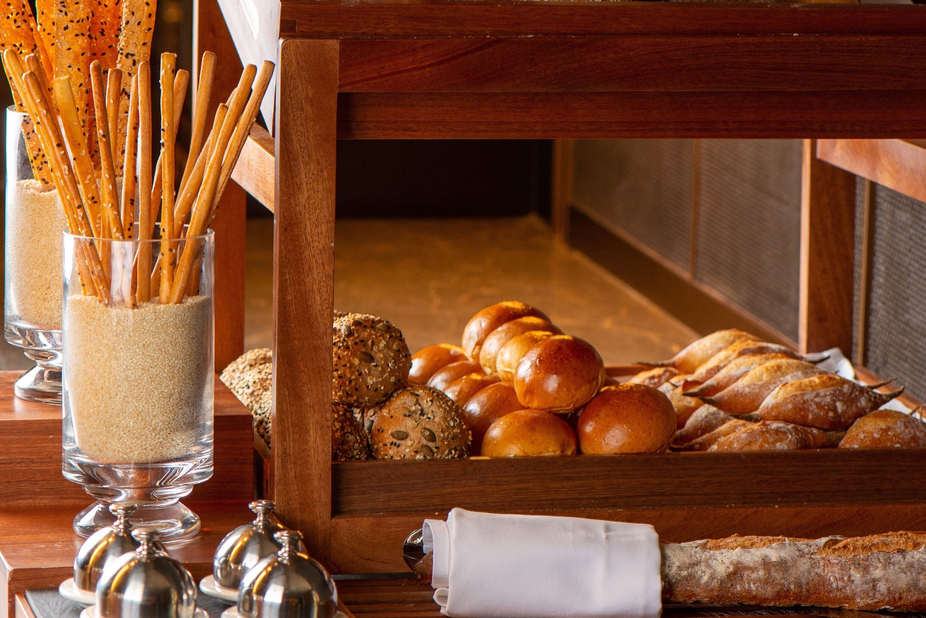 A buffet table with various breads and rolls, a glass container with pretzel sticks, and a napkin.