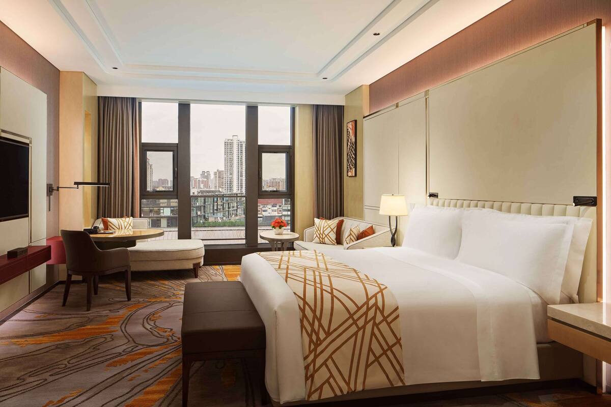 A hotel room with a large bed, a desk, a chair, a small table with a lamp, and a view of the city through large windows.