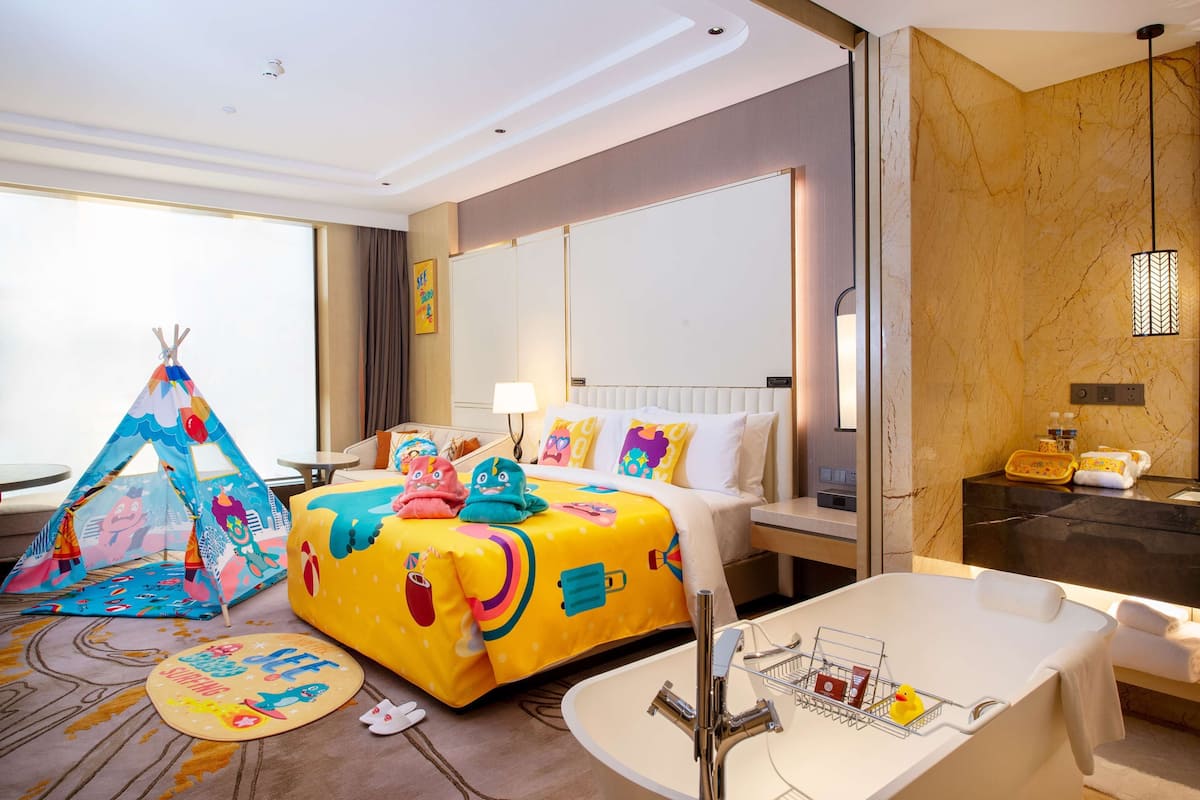 A hotel room with a colorful bed, a bathroom with a bathtub, a tent, and a large window.