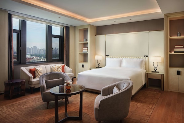Una habitación de hotel con una cama grande, un sofá, una silla, una mesita y vistas de la ciudad a través de amplias ventanas.