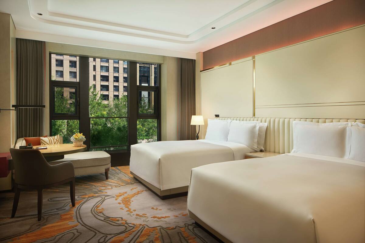A hotel room with two beds, a desk, a chair, a window with a view of buildings, and a carpet with a geometric pattern.