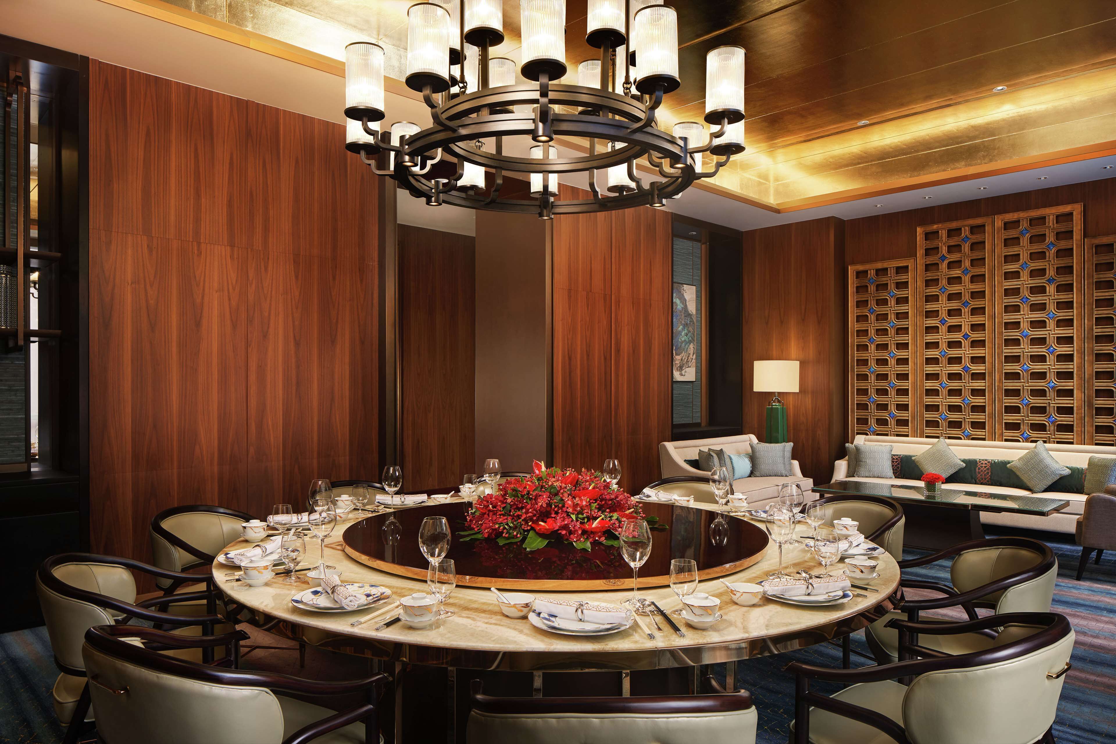 A round dining table with a floral centerpiece, surrounded by chairs, in a room with wooden paneling and a chandelier.