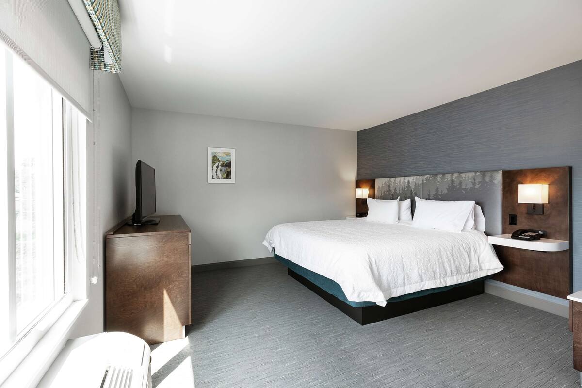 A modern hotel room with a large bed, a bedside table with a lamp, a flat-screen TV on a wooden cabinet, and a window with blinds.
