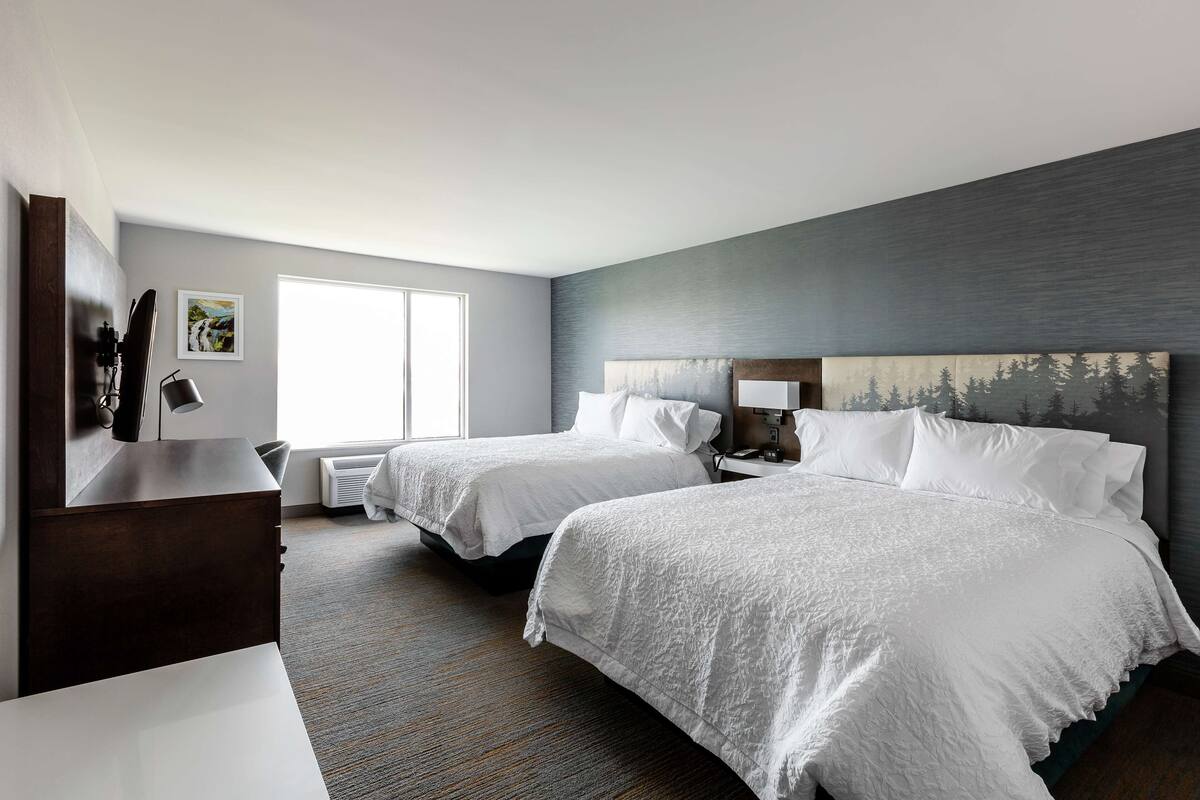 A hotel room with two beds, a desk, a chair, a window, and a painting on the wall.