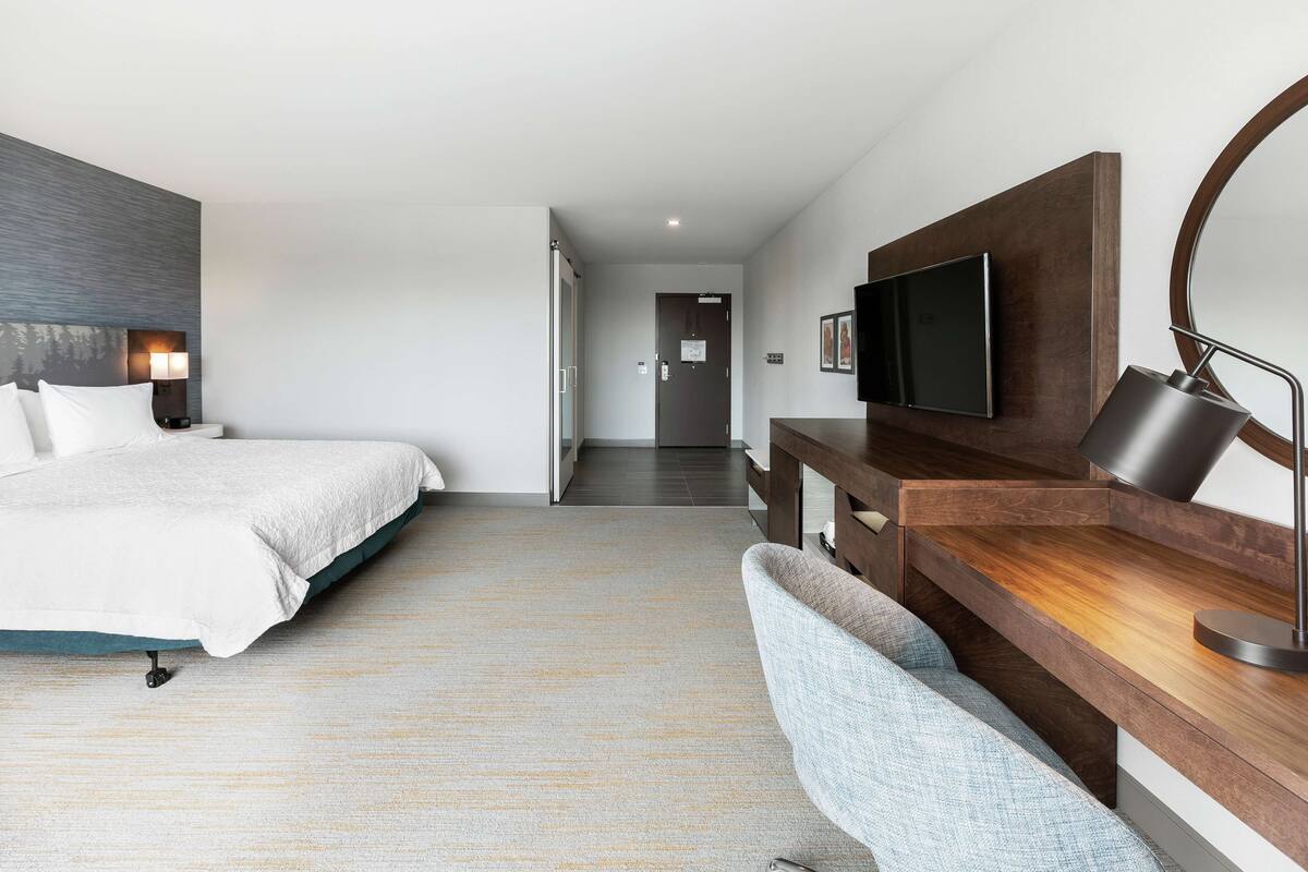 A modern hotel room with a bed, a desk with a computer, a chair, a TV, and a mirror.