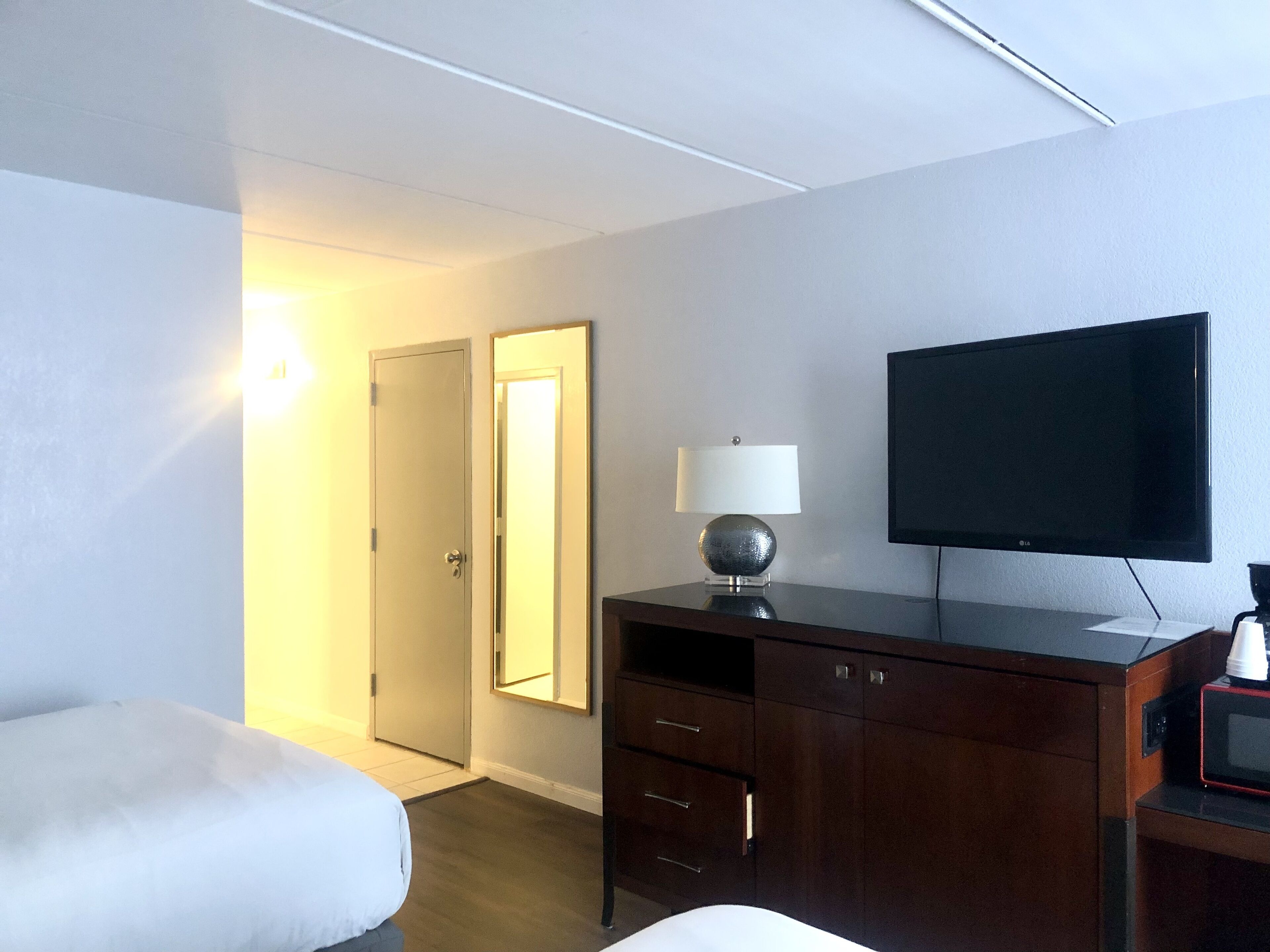 A hotel room with a bed, a desk with a lamp, a television, and a door.