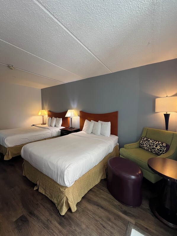 Una habitación de hotel con dos camas, un sillón verde, una mesita pequeña y un reposapiés morado.
