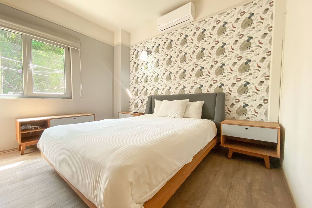 Een slaapkamer met een groot bed, twee nachtkastjes, een wandairconditioner en een raam met uitzicht op groen.