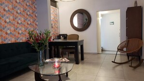 Een moderne woonkamer met een glazen salontafel, een donkergroene bank, een eettafel met stoelen, een rieten schommelstoel en een wand met een mozaïek van honingraat tegels.