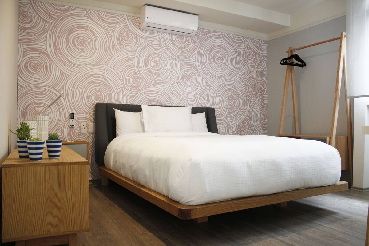 Een slaapkamer met een bed, een houten nachtkastje, een wandairconditioner en een houten kledingrek.