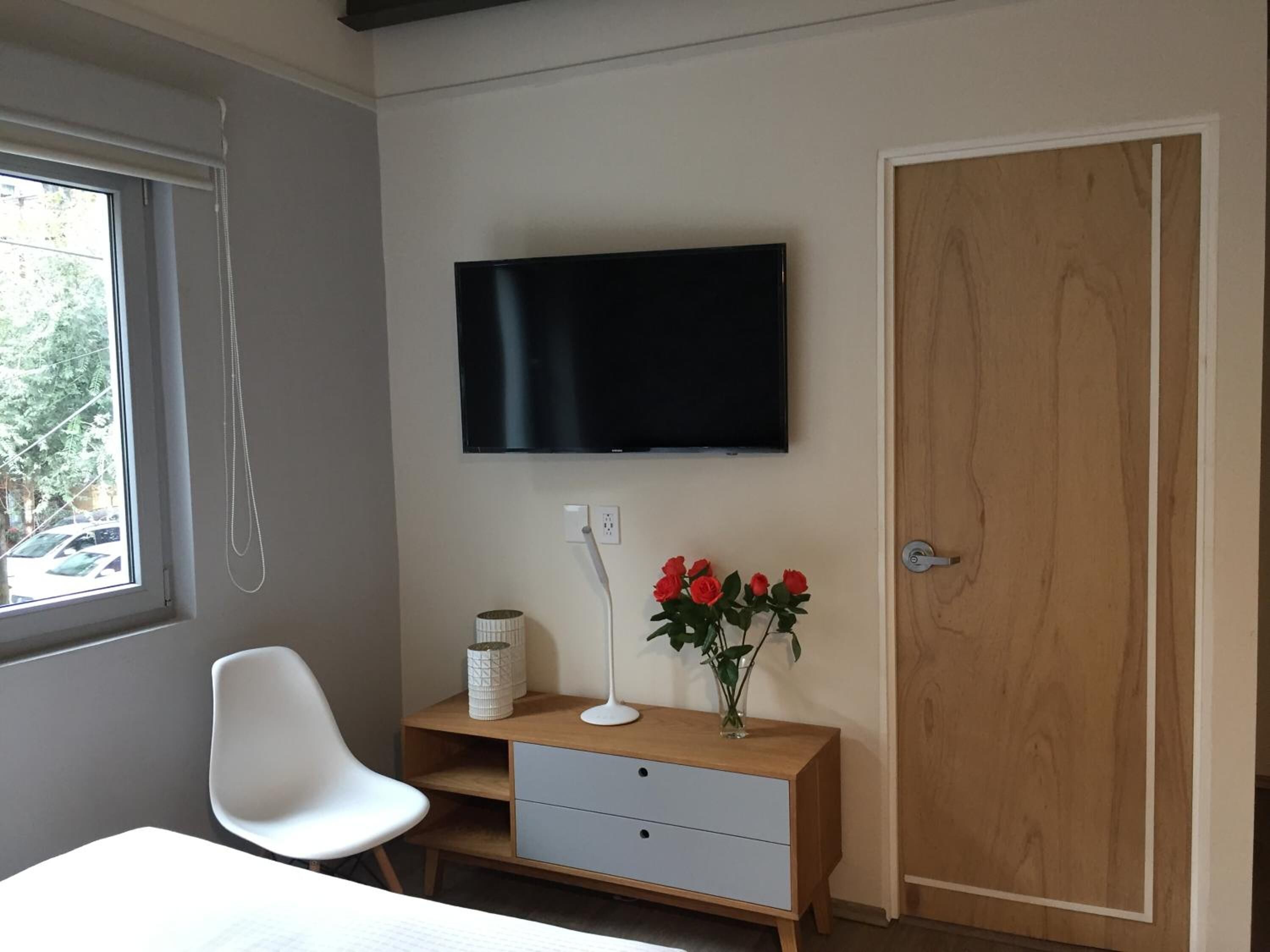 Un dormitorio con cama, un escritorio con lámpara, un televisor montado en la pared y una puerta de madera.