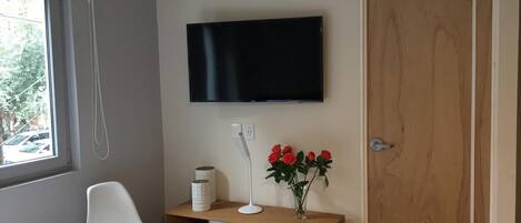 Un dormitorio con cama, un escritorio con lámpara, un televisor montado en la pared y una puerta de madera.