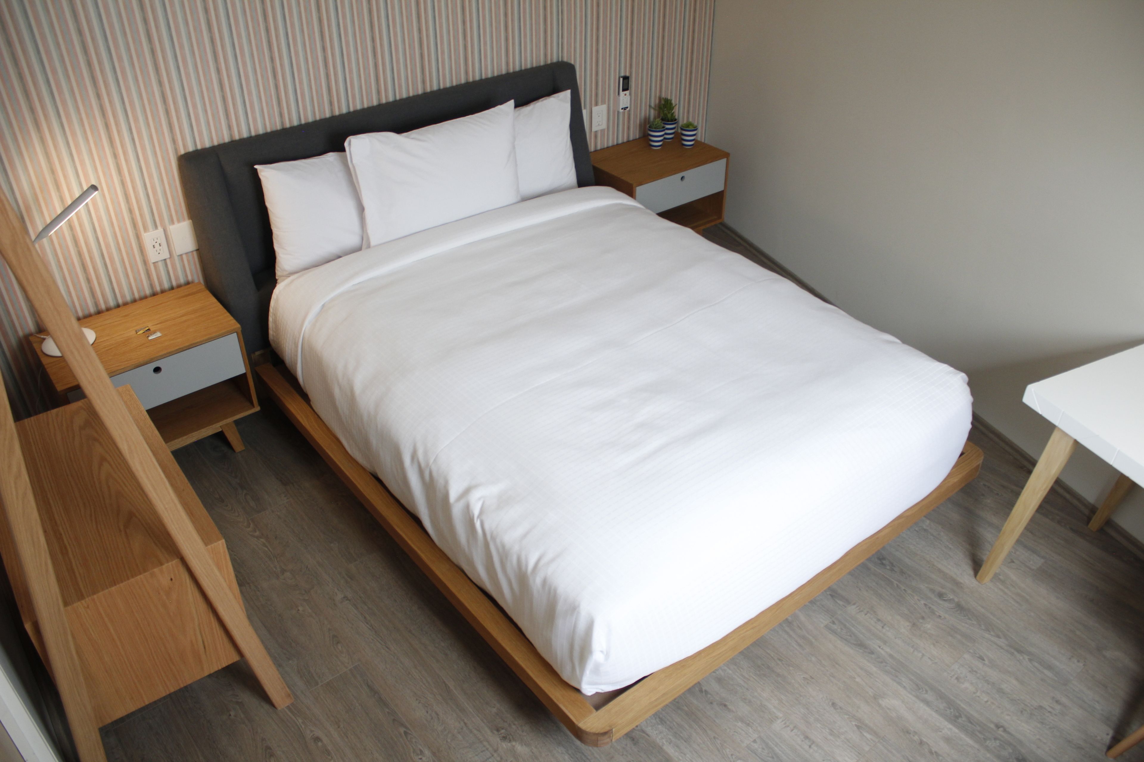 A neatly made bed with white linens, a wooden headboard, and a nightstand with a lamp and a small potted plant.