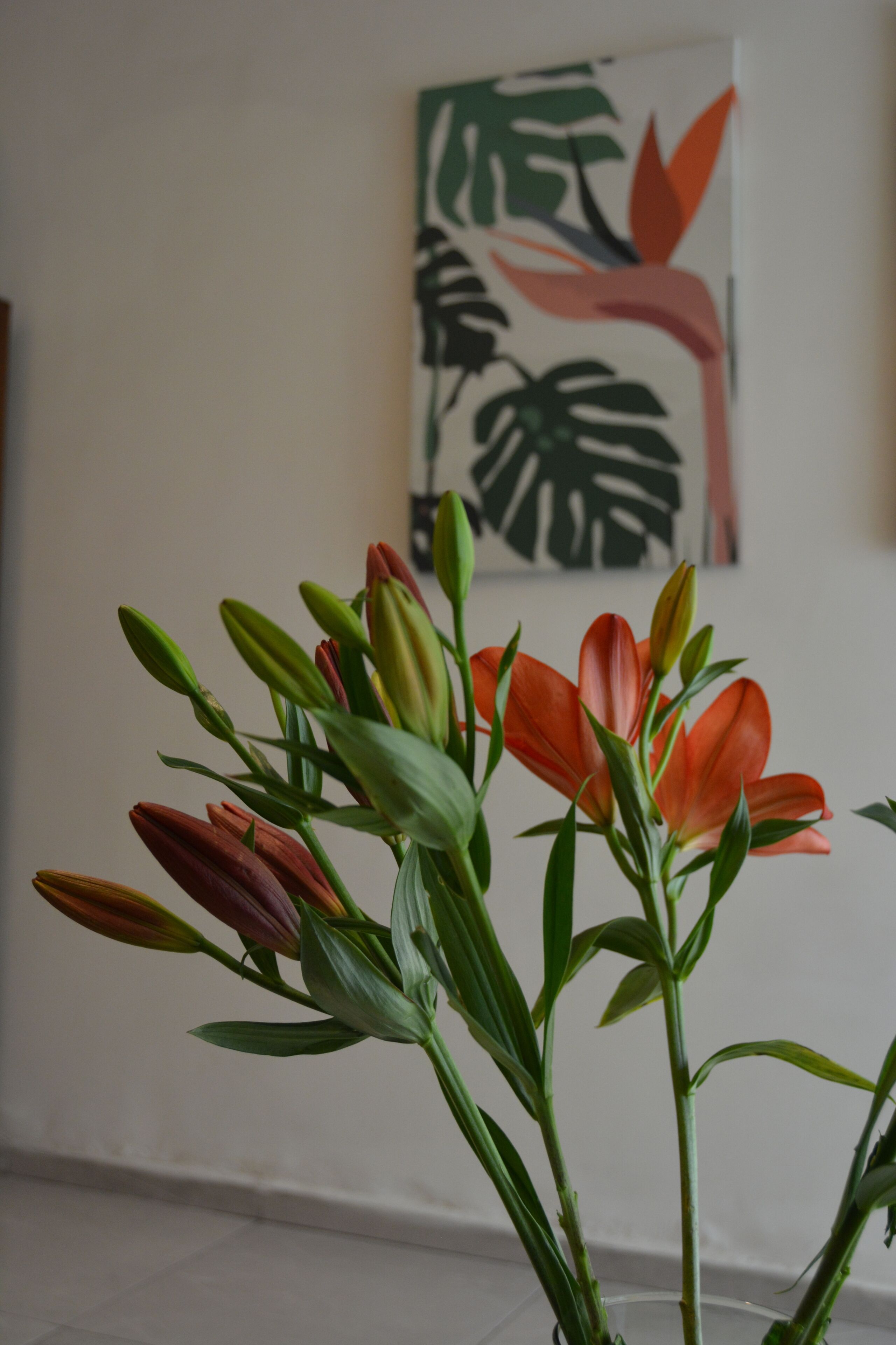 A room with a vase of flowers on a table, a painting on the wall, and a white tiled floor.
