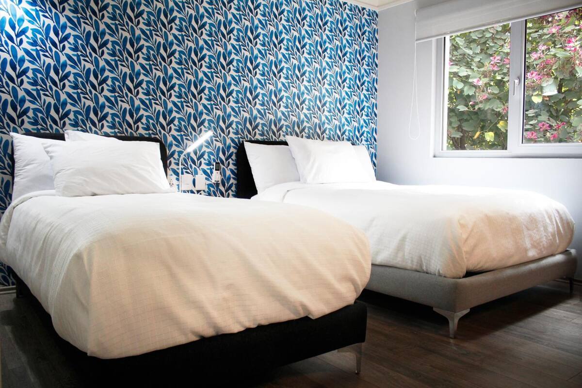 Een tweepersoonsbed met een zwart frame en witte beddengoed, een eenpersoonsbed met een grijs frame en witte beddengoed, en behang met een blauw bladpatroon.