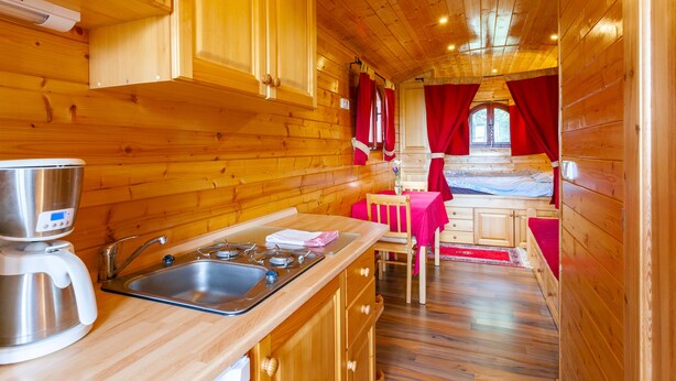 L’intérieur d’un chalet en bois comprend un coin cuisine, une table à manger avec des rideaux rouges et un lit recouvert d’une couverture rouge.