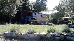 Une cabane rustique avec un toit bleu et des murs en bois, entourée d’un mur de pierres et d’une zone herbeuse.