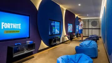 Un espace de jeux vidéo doté de plusieurs écrans diffusant des jeux, de fauteuils-pouf pour s’asseoir et d’une personne utilisant une tablette.