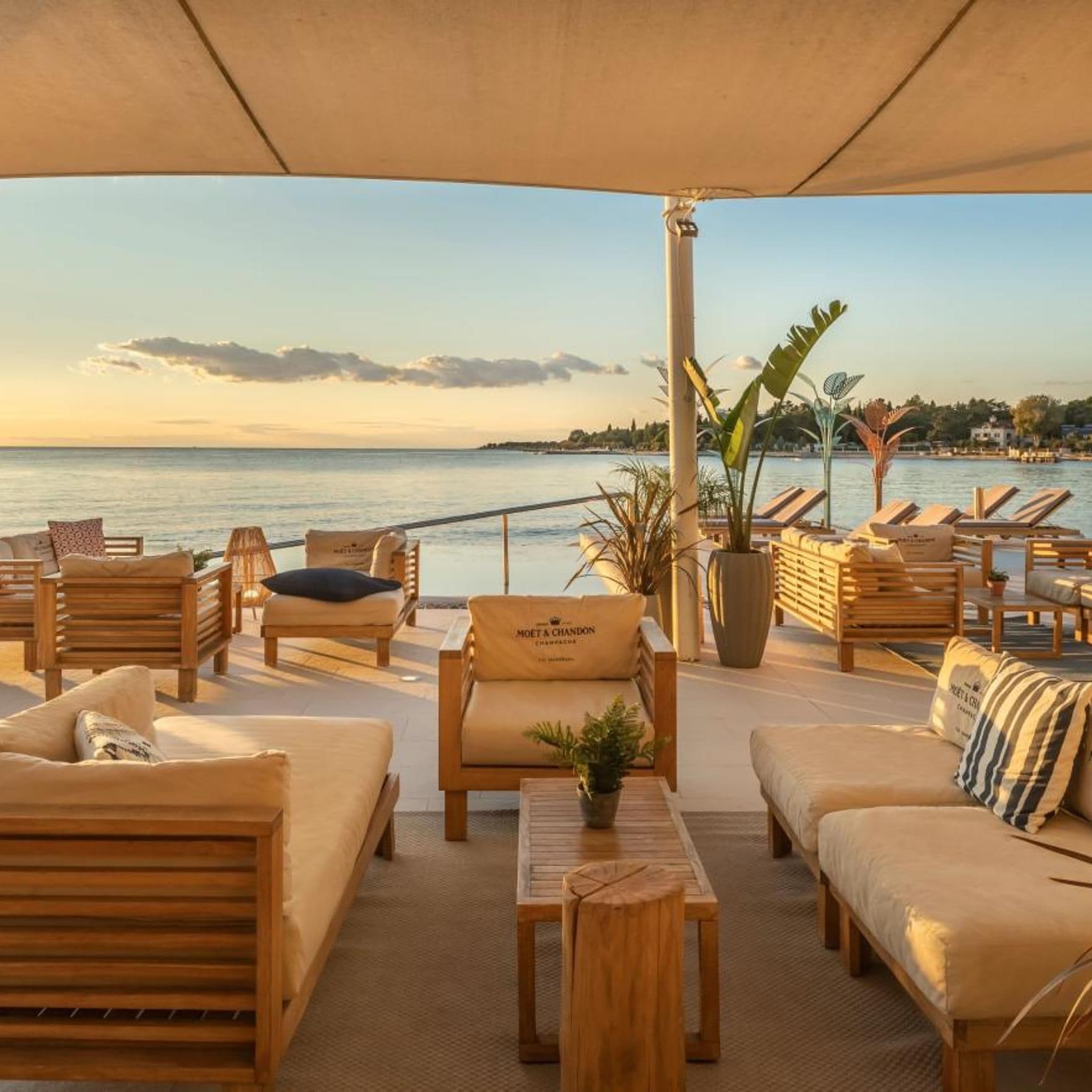 Ein sonniger Außenbereich mit Liegestühlen und Sofas, einem Holztisch und Blick auf das Meer.