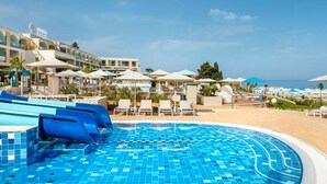 A hotel pool area with a clear sky, lounge chairs, umbrellas, and a building with a sign. - Marea Valamar Collection Suites (Porec)