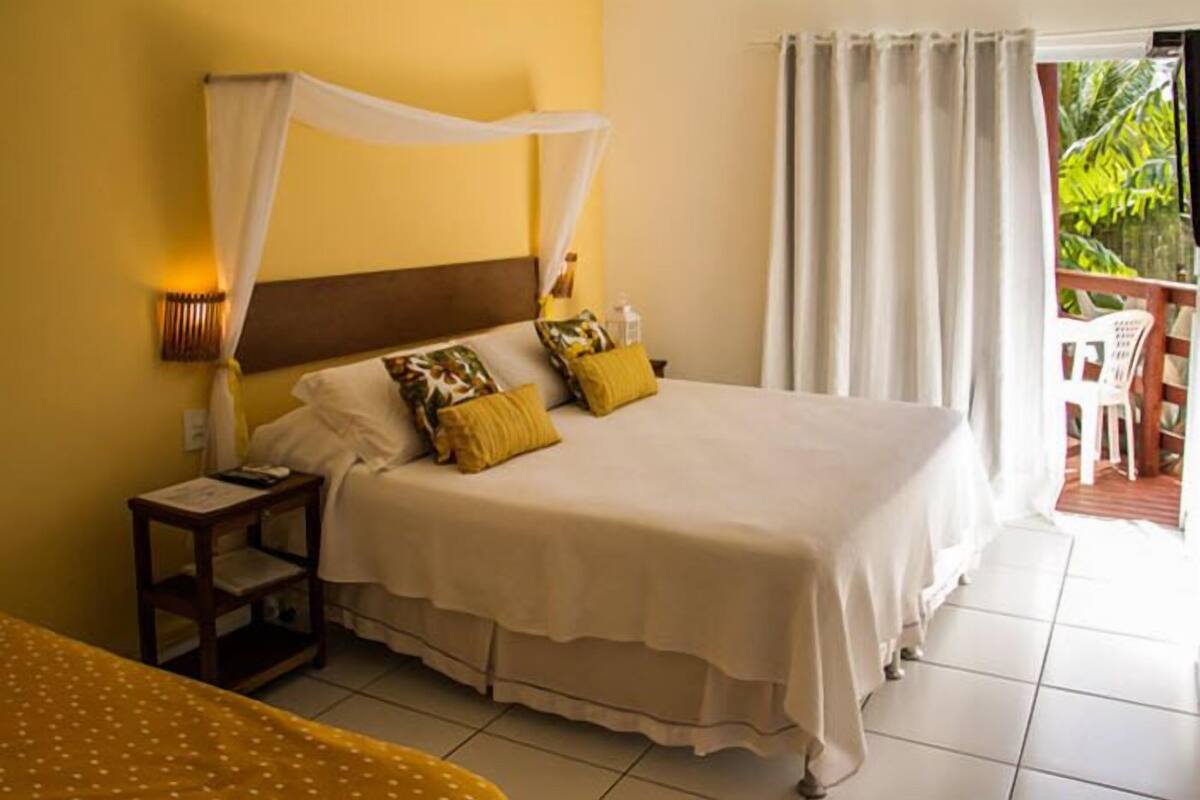 A bed with a canopy, a bedside table, and a chair in a room with yellow walls and a view of greenery outside.