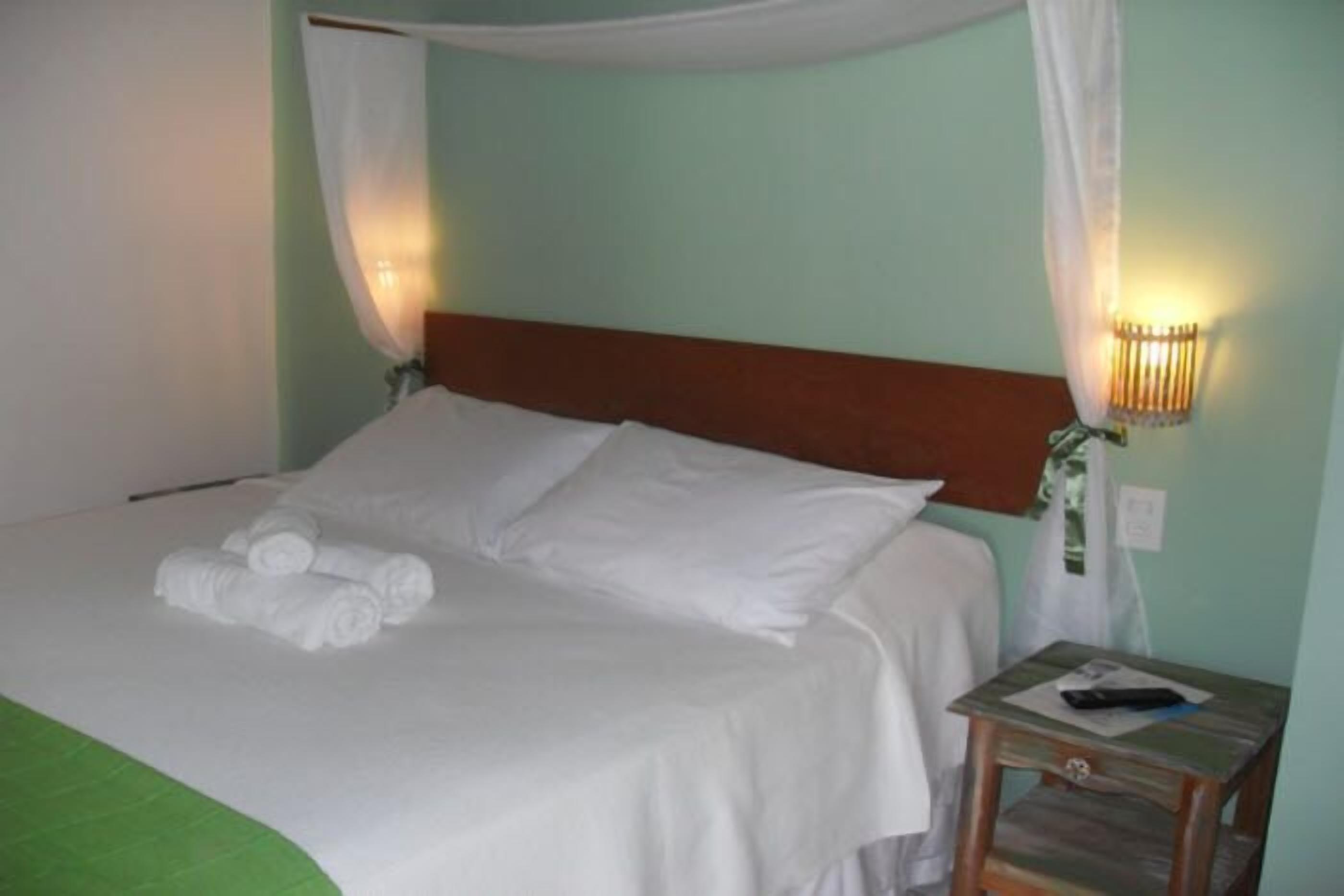 Una cama bien tendida con sábanas blancas y una colcha verde, una mesita de noche con una lámpara, y un aplique en la pared.