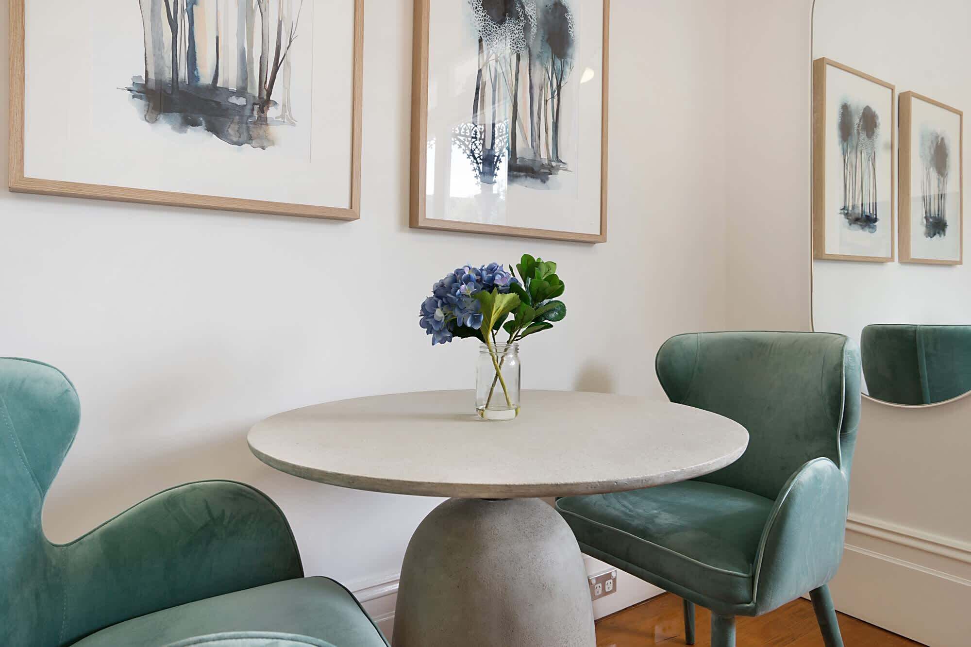 A dining area with a round table, two teal chairs, and a vase with blue flowers.