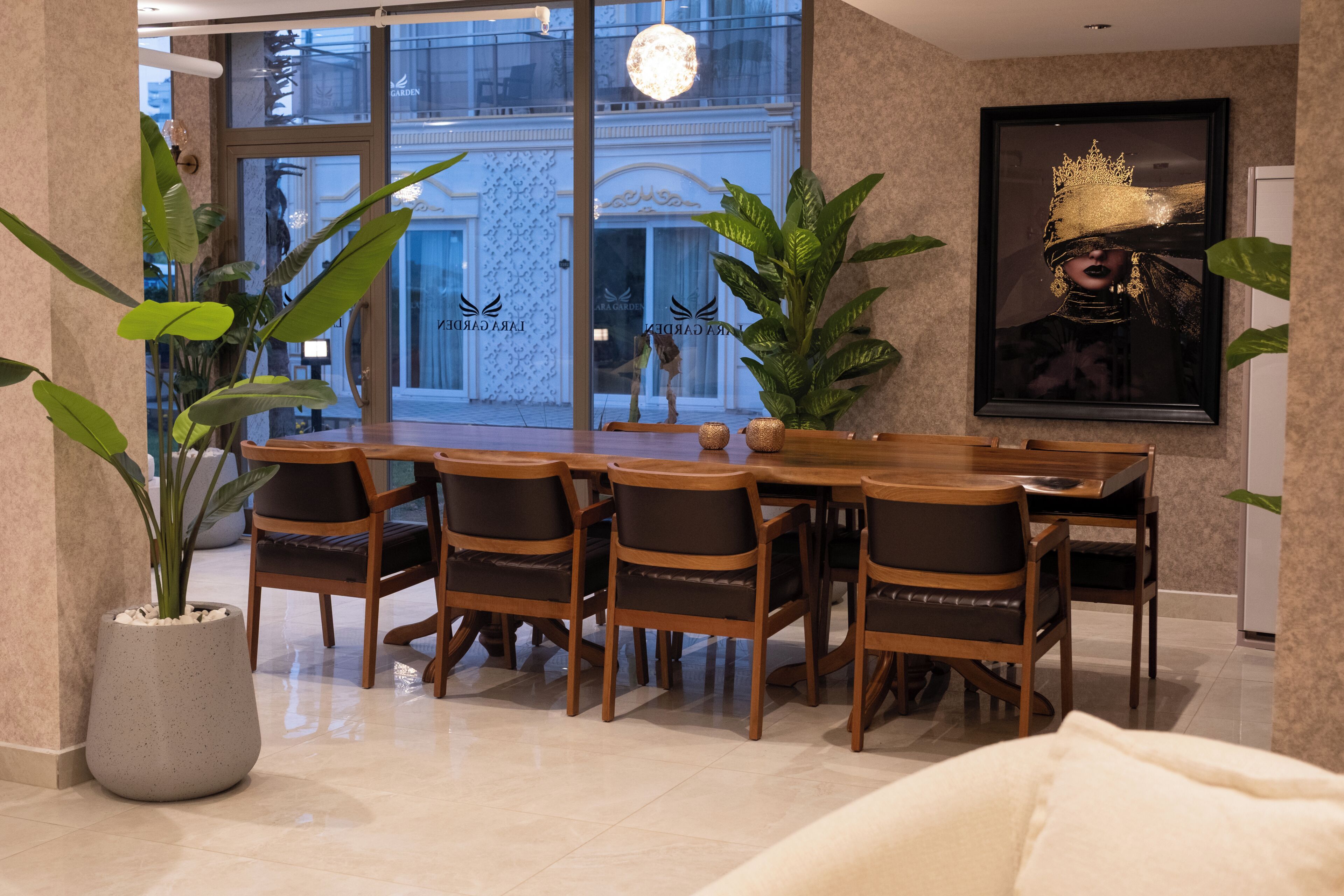 A dining area with a wooden table and chairs, potted plants, and a large window with a view of a building.