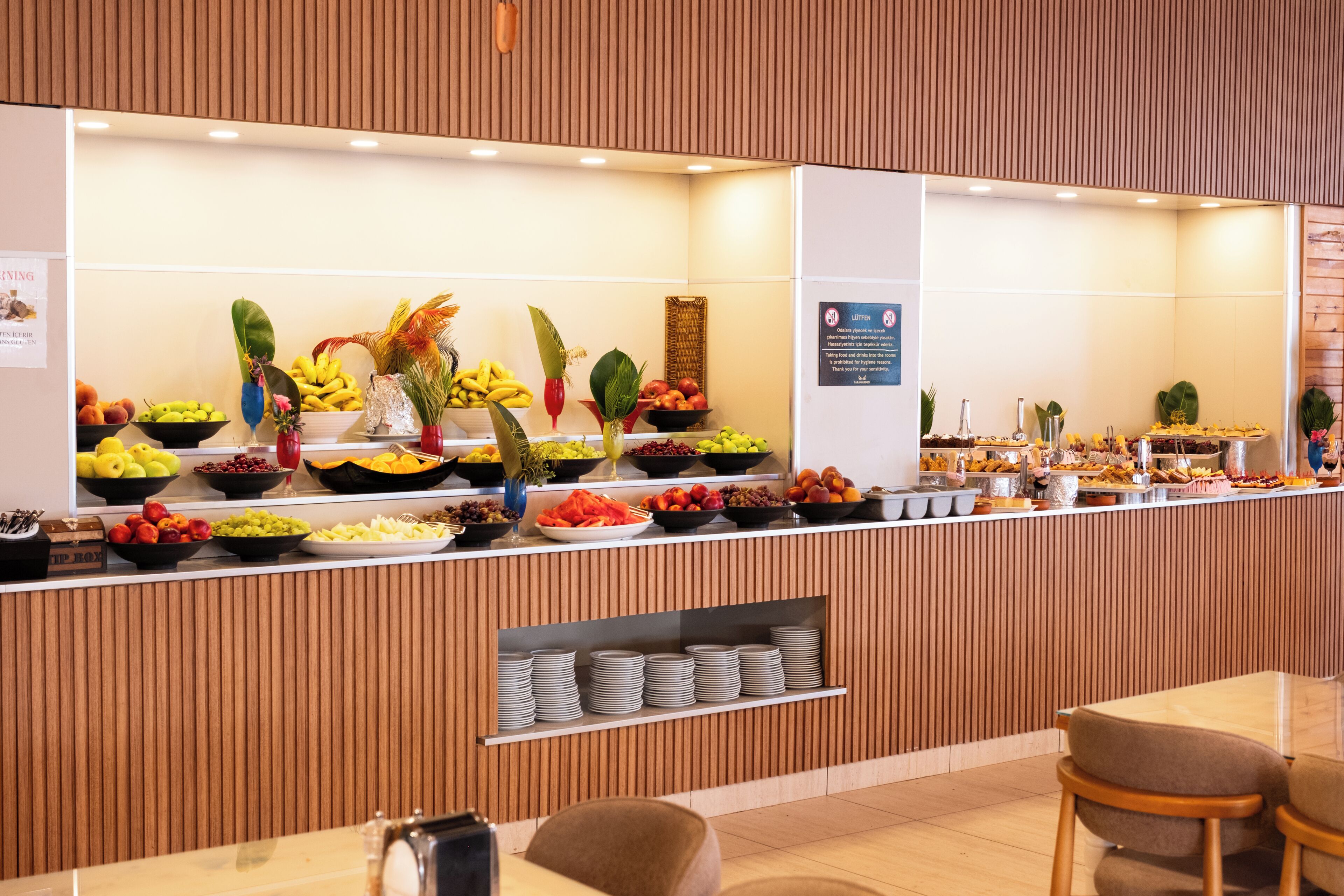 A buffet counter with various fruits and vegetables, a sign prohibiting food and drinks in the room, and a stack of plates.