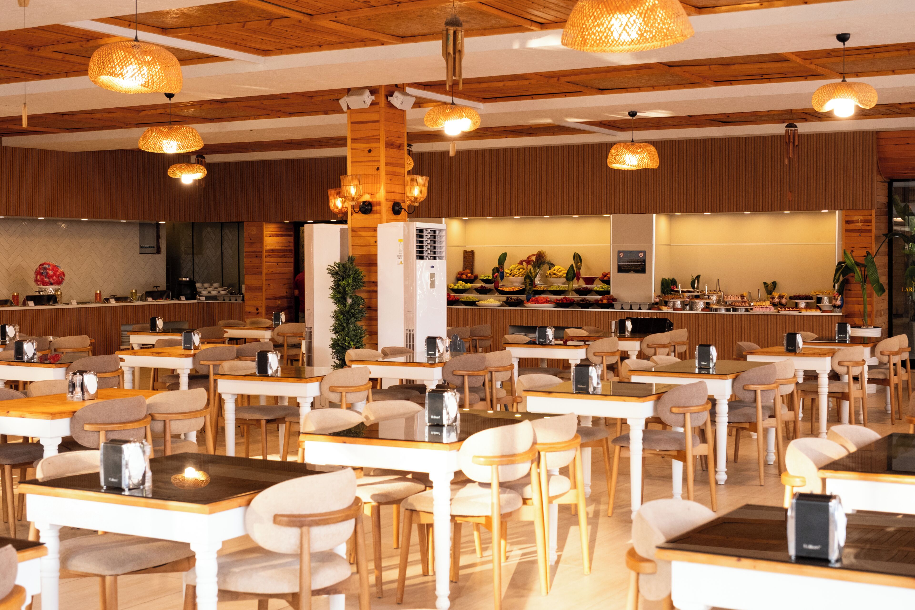 Un restaurante con mesas y sillas de madera, luces colgantes y una zona de bufé al fondo.